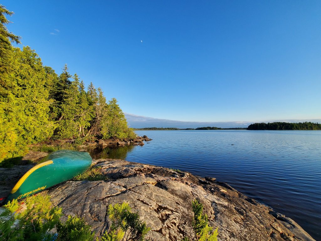 An alternative to Ontario crown land camping is using parks that have backcountry permit systems. Imagine shows green canoe in front of a large lake with pines trees.